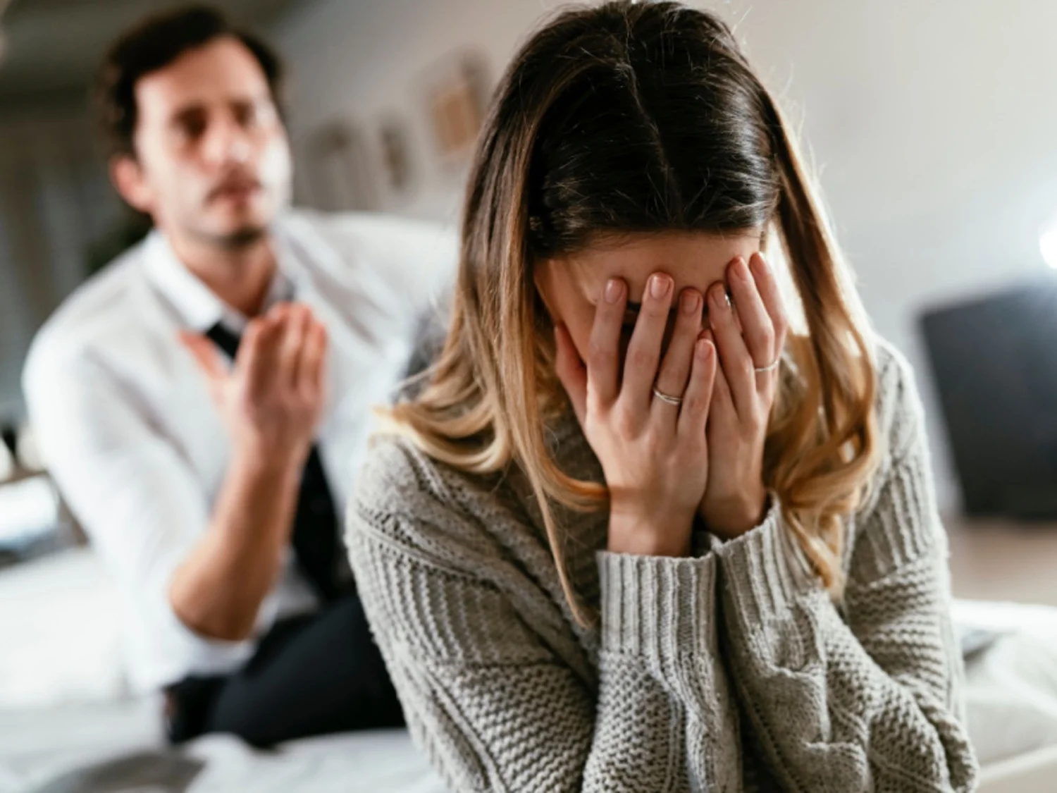 Man in background gestures angrily at upset woman with hands on her face