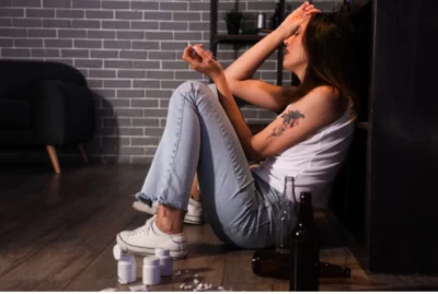 Young woman who is addicted to pills and alcohol sits on the floor looking distressed