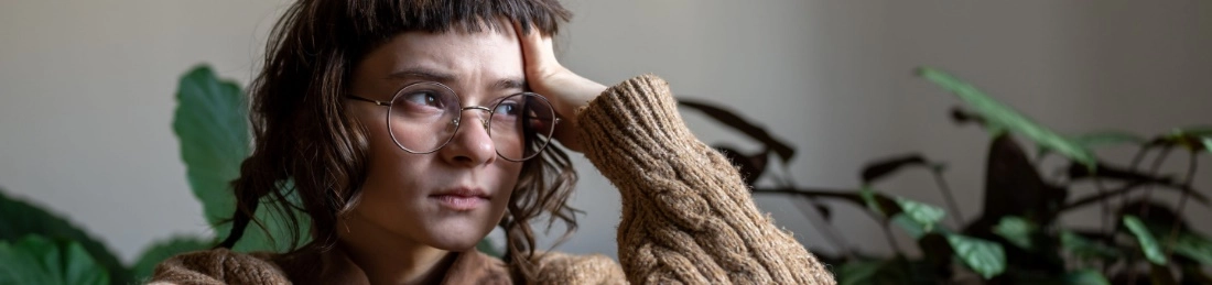 Young woman with glasses and shaggy haircut looks somberly out the window