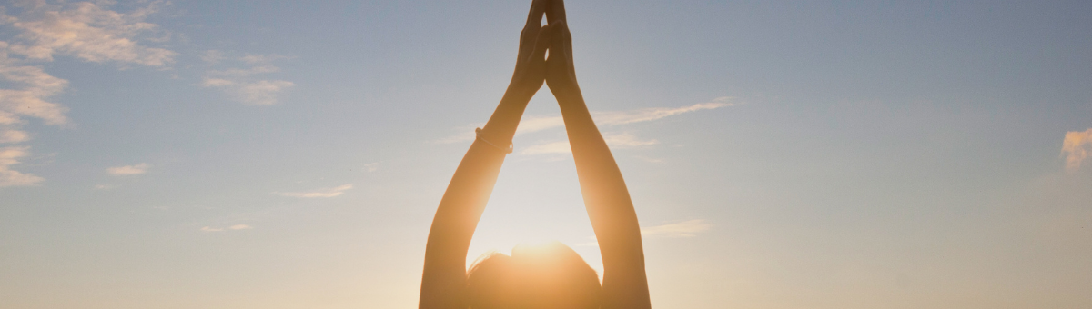 A woman practicing yoga with her hands raised in a Namaste pose, silhouetted against a vibrant sunset with a clear sky.