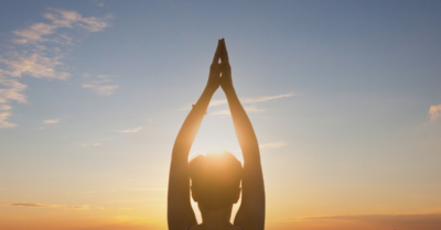 A woman practicing yoga with her hands raised in a Namaste pose, silhouetted against a vibrant sunset with a clear sky.